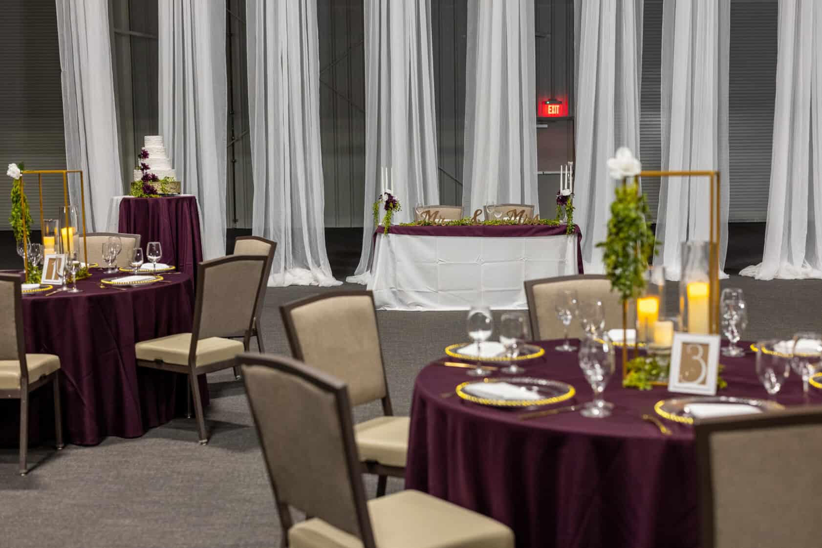 Tables with purple tablecloths, formal place settings, and candles are arranged in front of a white-draped backdrop and a decorated sweetheart table with a wedding cake in the background.