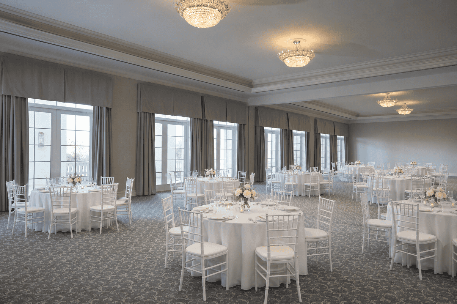Spacious banquet hall with round tables covered in white linens, white chairs, floral centerpieces, large windows, and chandeliers.