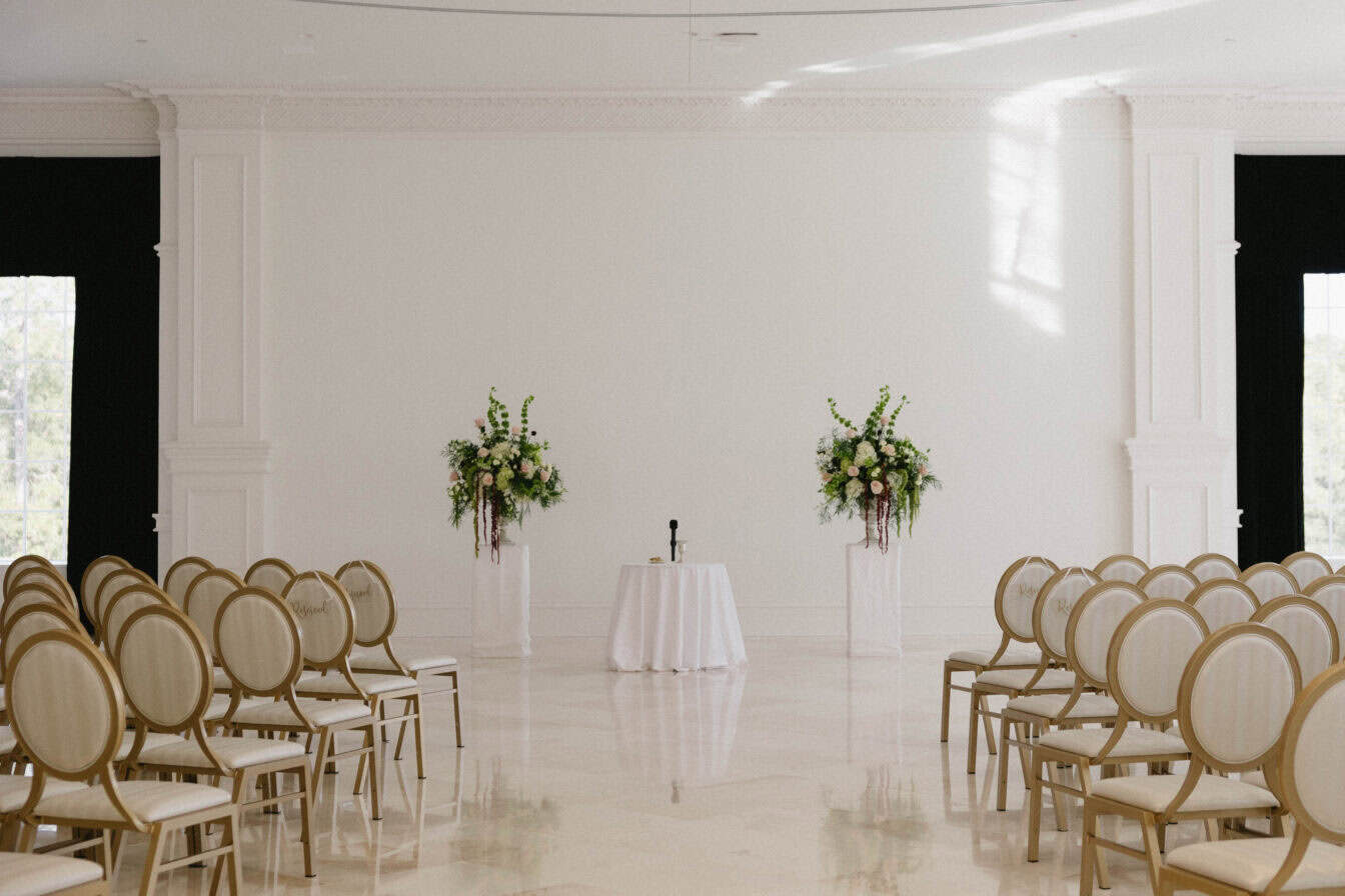 Rows of empty chairs face a small round table with a microphone, flanked by two tall floral arrangements in a bright, elegant room.