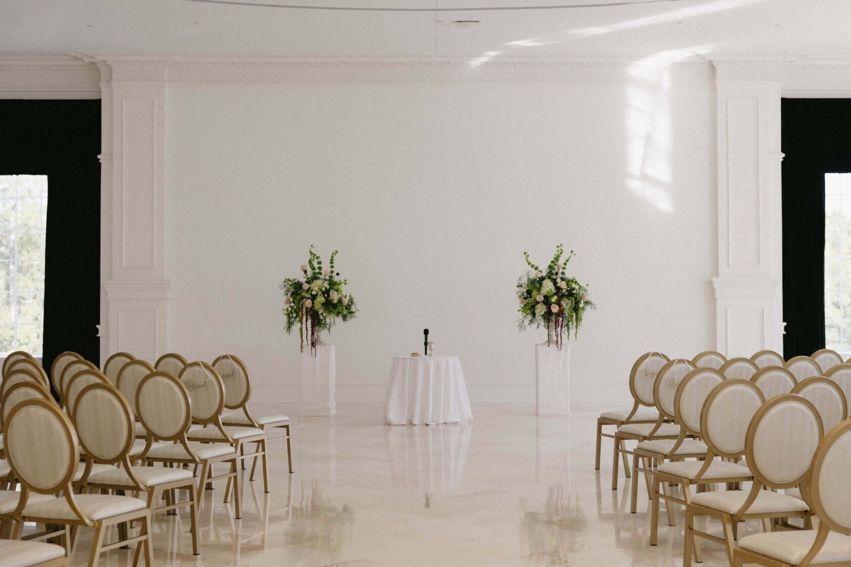 Rows of empty chairs face a small round table with a microphone, flanked by two tall floral arrangements in a bright, elegant room.