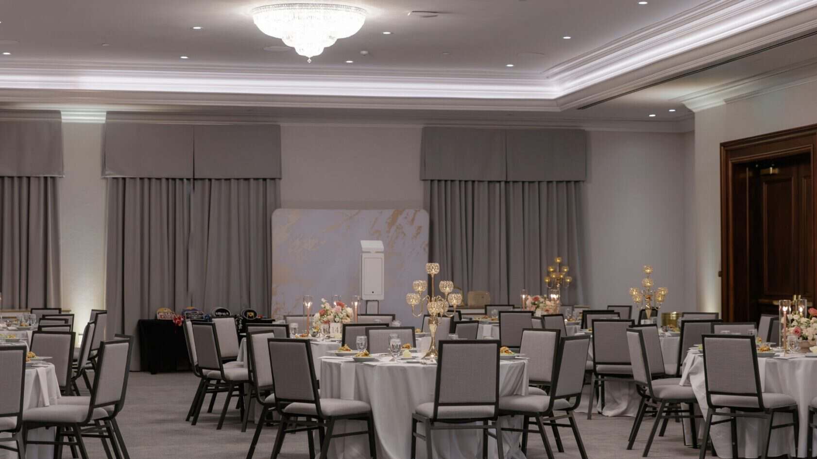 Banquet hall with round tables set for an event, featuring neutral decor, floral centerpieces, and gray chairs under a chandelier.