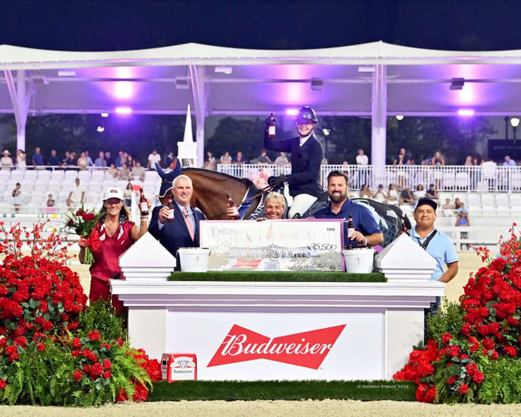 A group of people, including a person on horseback, pose behind a large check and Budweiser sign at an outdoor event, surrounded by red flowers and spectators in the background.