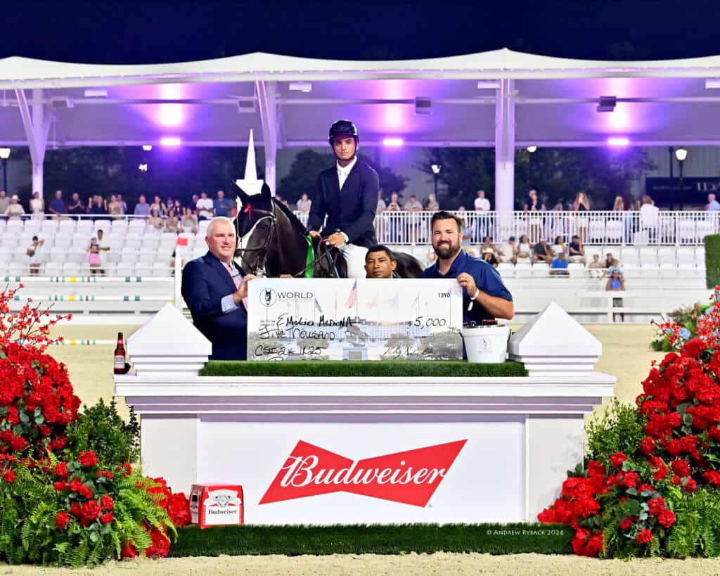 A group of people poses with a horse and an oversized $5,000 check at an outdoor equestrian event, standing behind a Budweiser-branded podium decorated with red flowers.