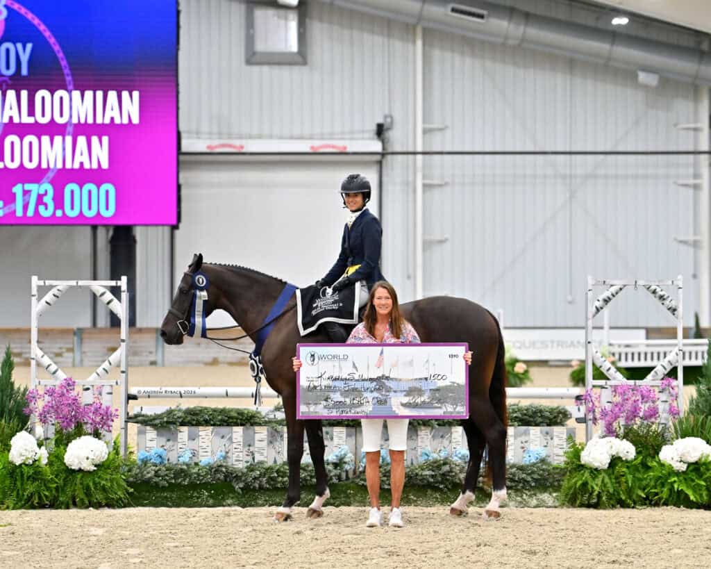A person in equestrian attire sits on a horse while another person stands in front holding a large check; flowers and a digital scoreboard are in the background.