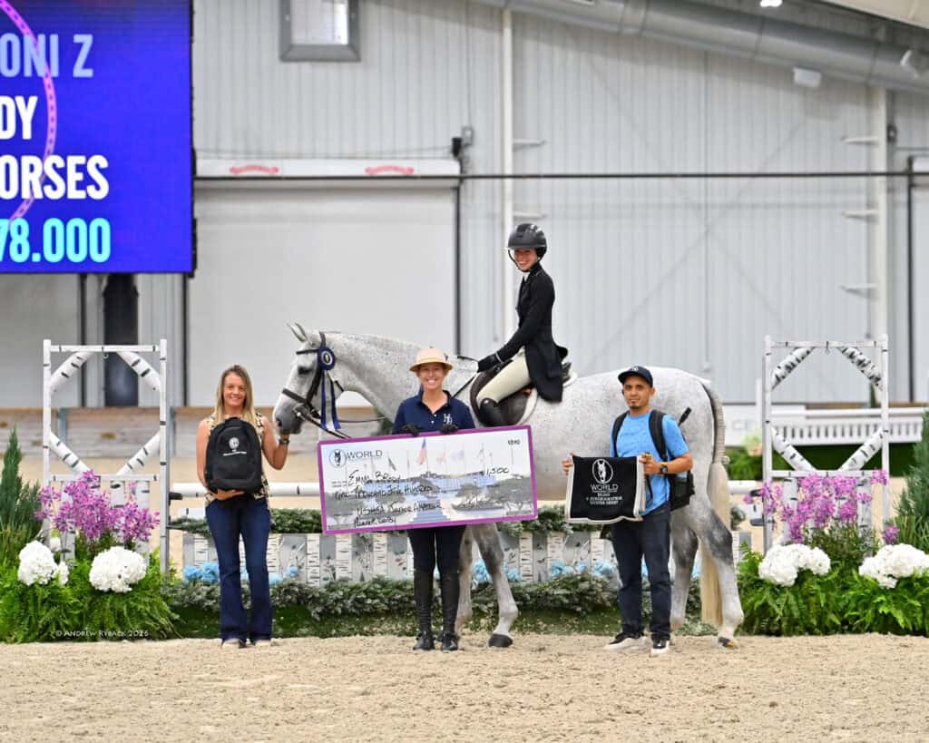 Four people and a horse pose indoors; one person is holding a large check, another sits on the horse, and banners and flowers decorate the arena.