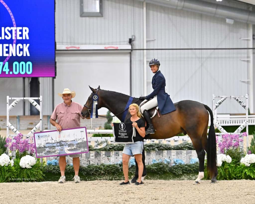 A rider on a horse poses indoors with two people; one person holds a large check and the other holds a trophy bag. Decorative flowers are in the background.