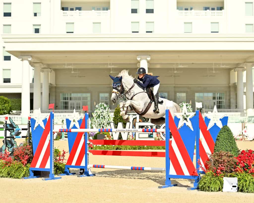 A rider on a white horse jumps over a colorful red, white, and blue obstacle during an equestrian event in front of a large building.