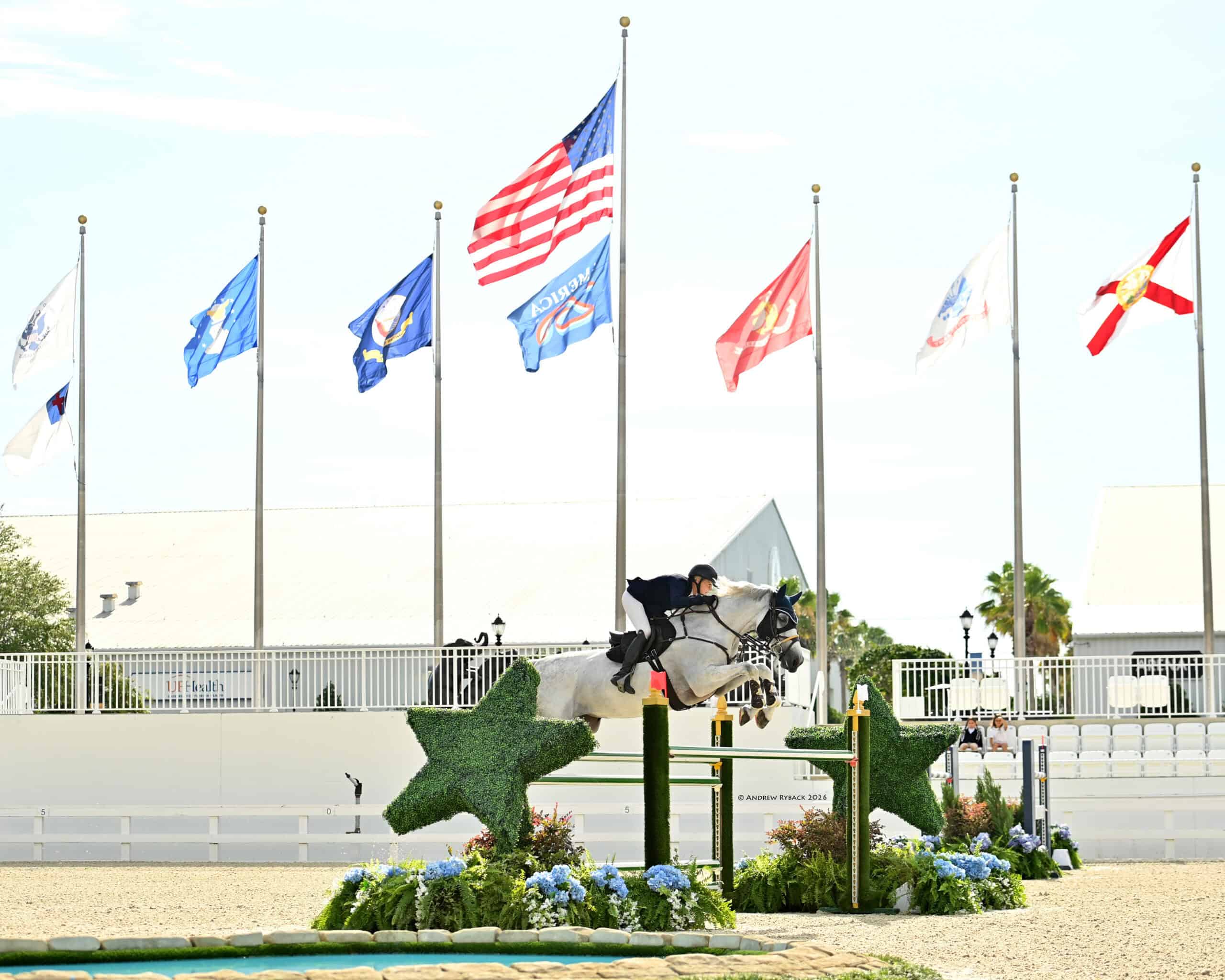 A rider on a white horse jumps over a green star-shaped obstacle in an outdoor arena, with several flags and a building in the background.
