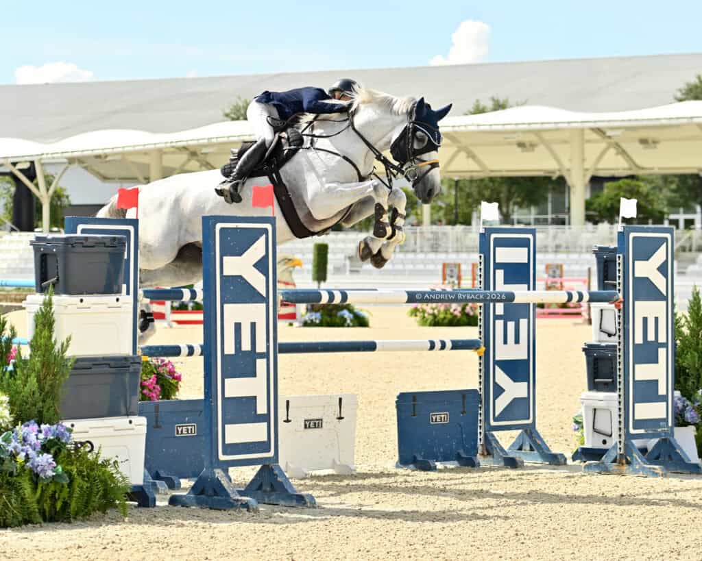 A rider on a white horse jumps over a YETI-branded obstacle during an equestrian show jumping competition in an outdoor arena.