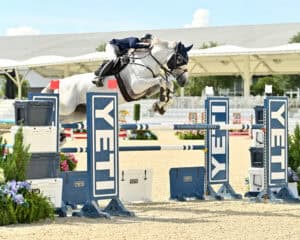 A rider on a white horse jumps over a YETI-branded obstacle during an equestrian show jumping competition in an outdoor arena.