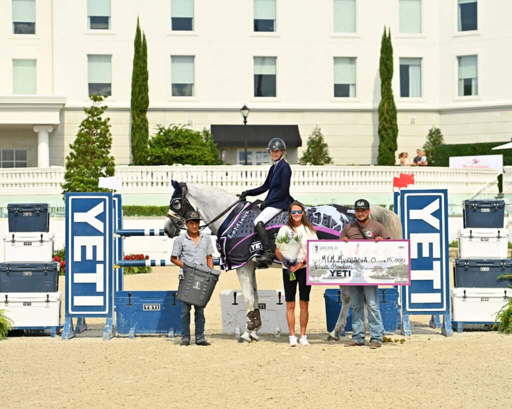 Four people and a horse pose at an equestrian event. One person holds a trophy, another holds a large $15,000 check. Event signage and YETI branding are visible.