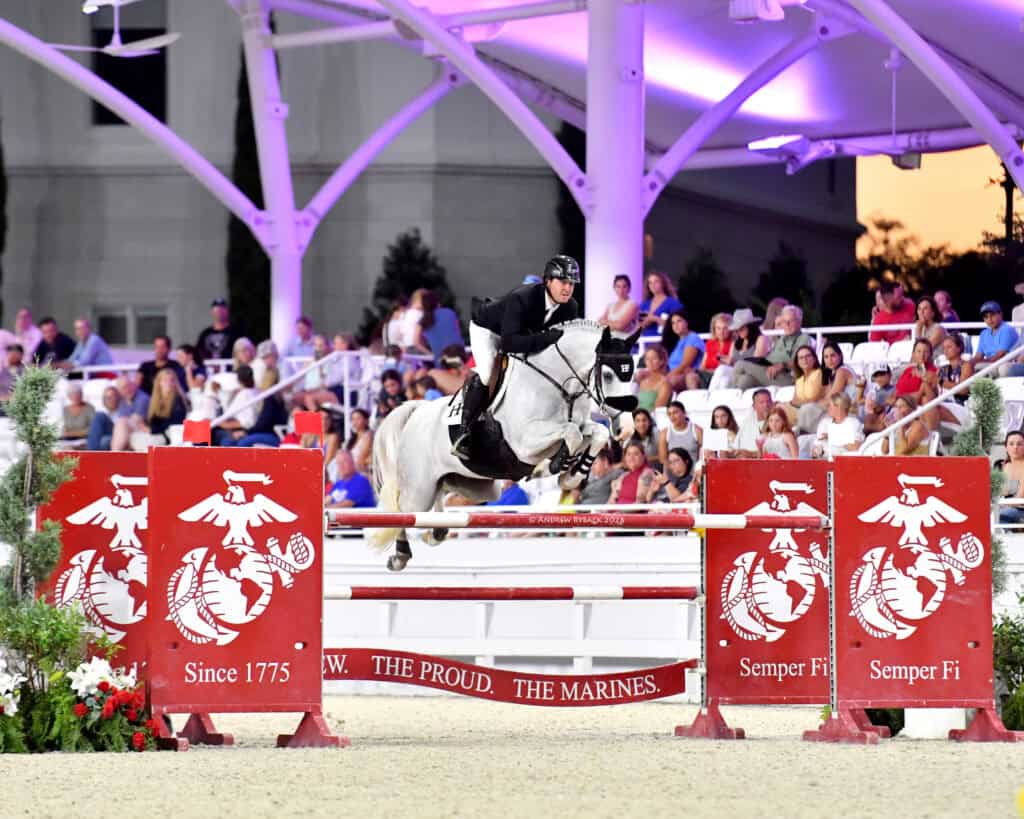 A rider on a white horse jumps over a red obstacle with Marine Corps logos during an equestrian event before a seated audience.