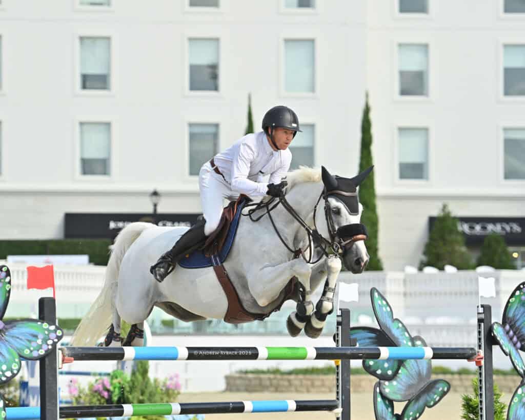 A rider in white attire and a black helmet guides a gray horse over a colorful jump during an equestrian event, with a building in the background.