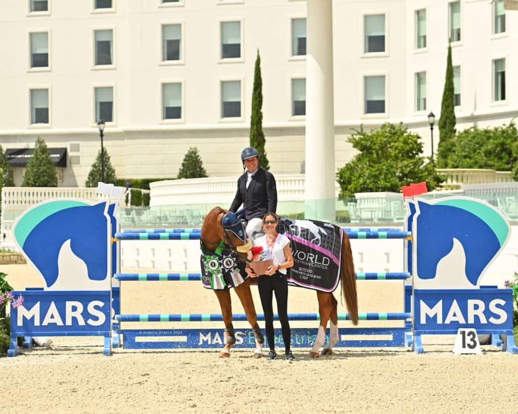 Equestrian rider on a horse, both wearing ribbons, poses with another person holding flowers in front of a jump with "MARS" branding at an outdoor event.