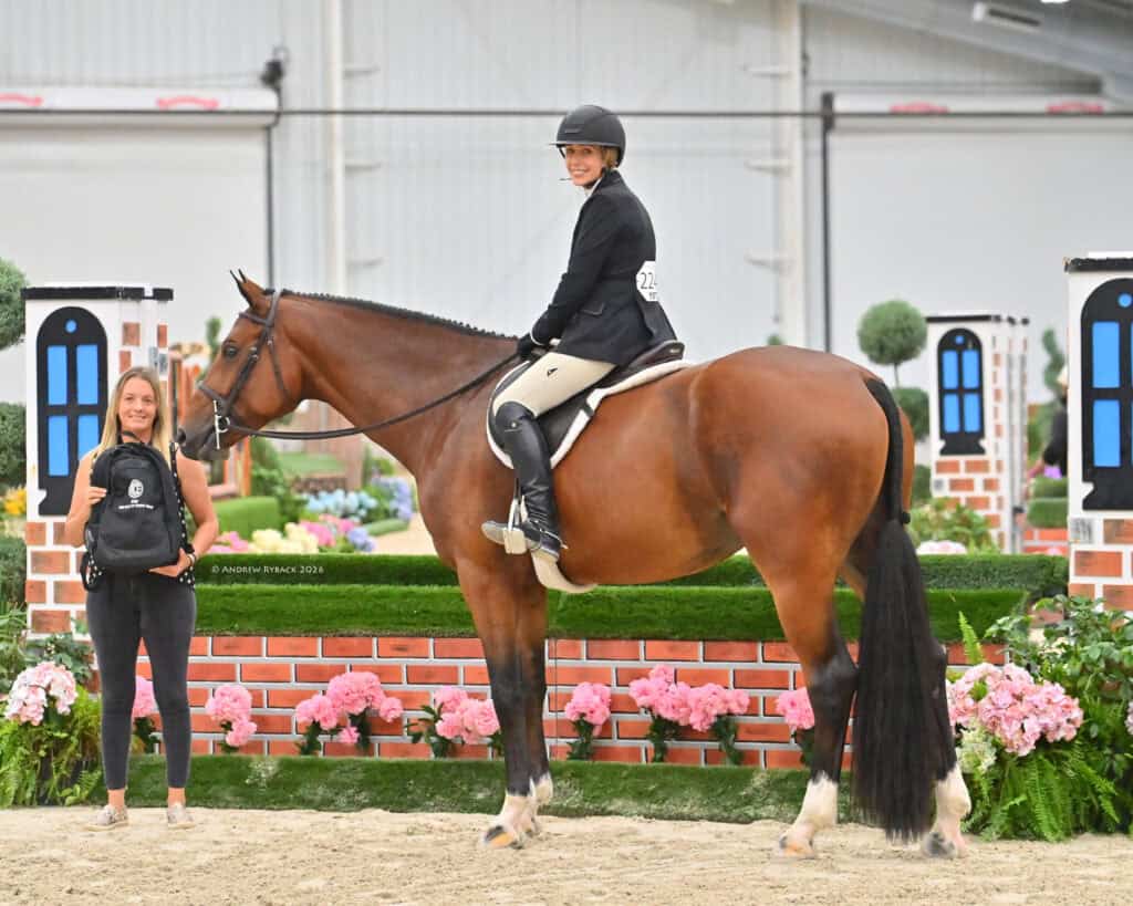 A rider in equestrian attire sits on a brown horse indoors, while a woman stands beside them holding a backpack. Decorative jumps and flowers are in the background.