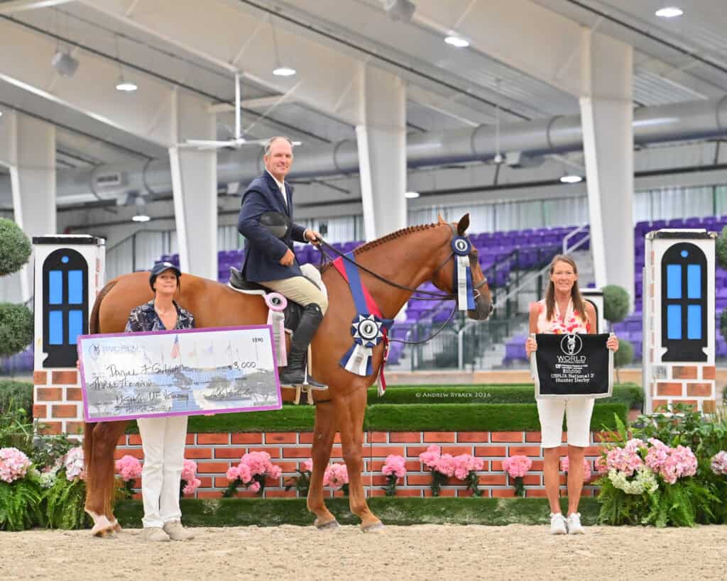 A man on a horse with a blue ribbon, flanked by two women holding a large check and a black sign, posing indoors with a brick and flower backdrop.