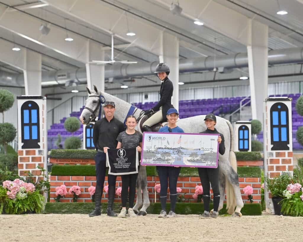 Five people and a horse pose indoors; one person holds a trophy banner, another holds an oversized check, and flowers decorate the area.