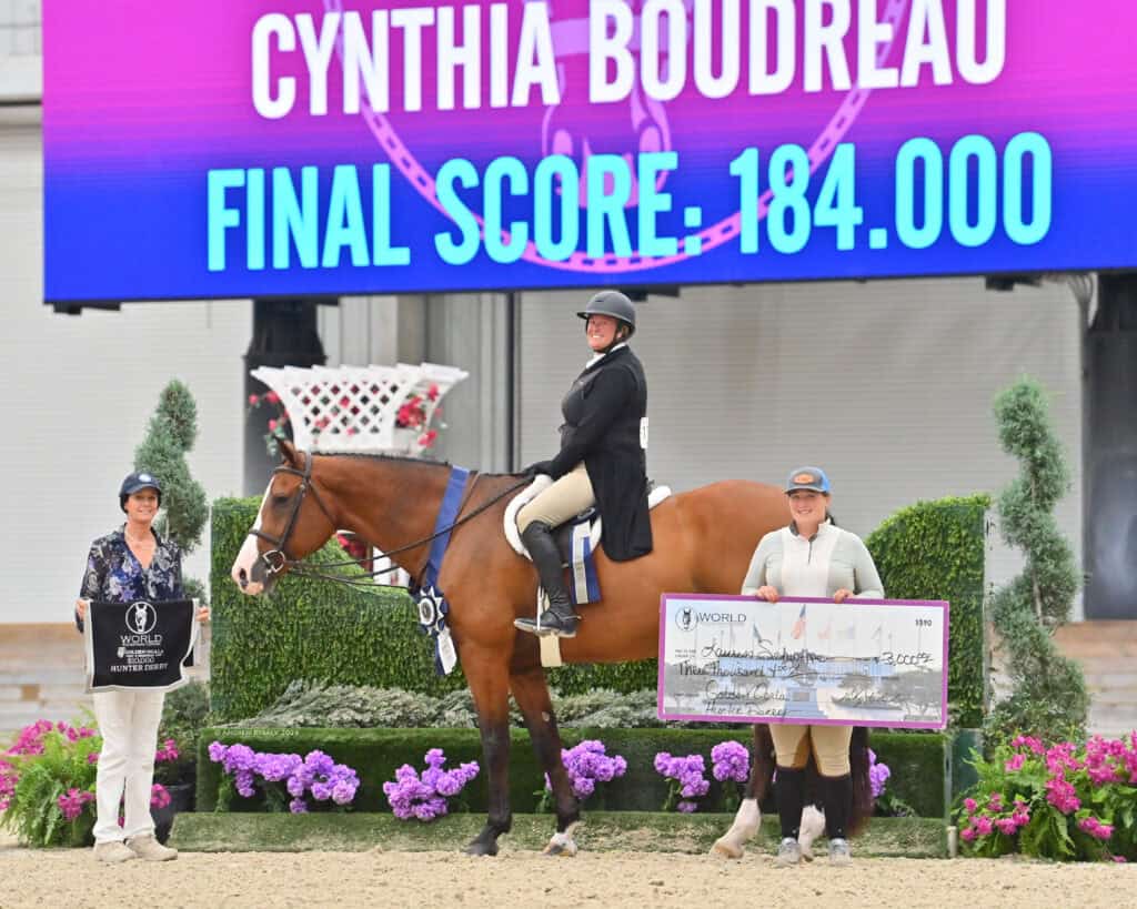 Equestrian rider on horse with two people beside her, one holding a trophy and one holding a large check, with event scoreboard displaying final score in the background.