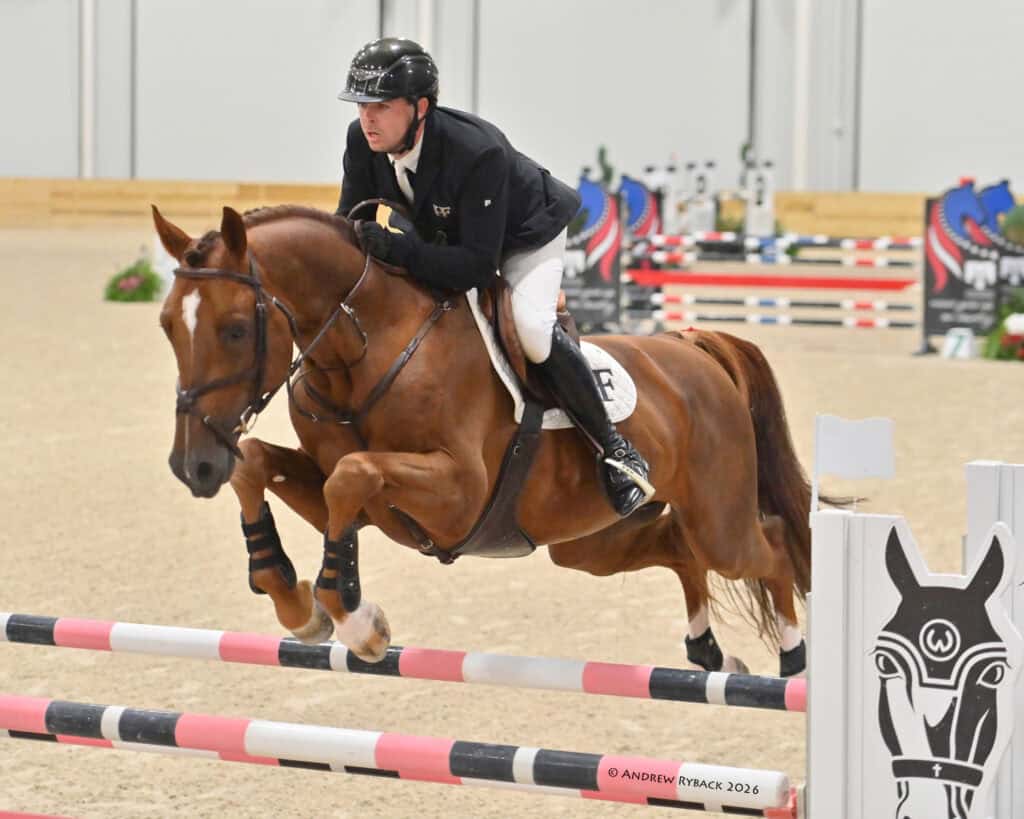A rider in equestrian attire guides a brown horse over a red, black, and white jump in an indoor arena.