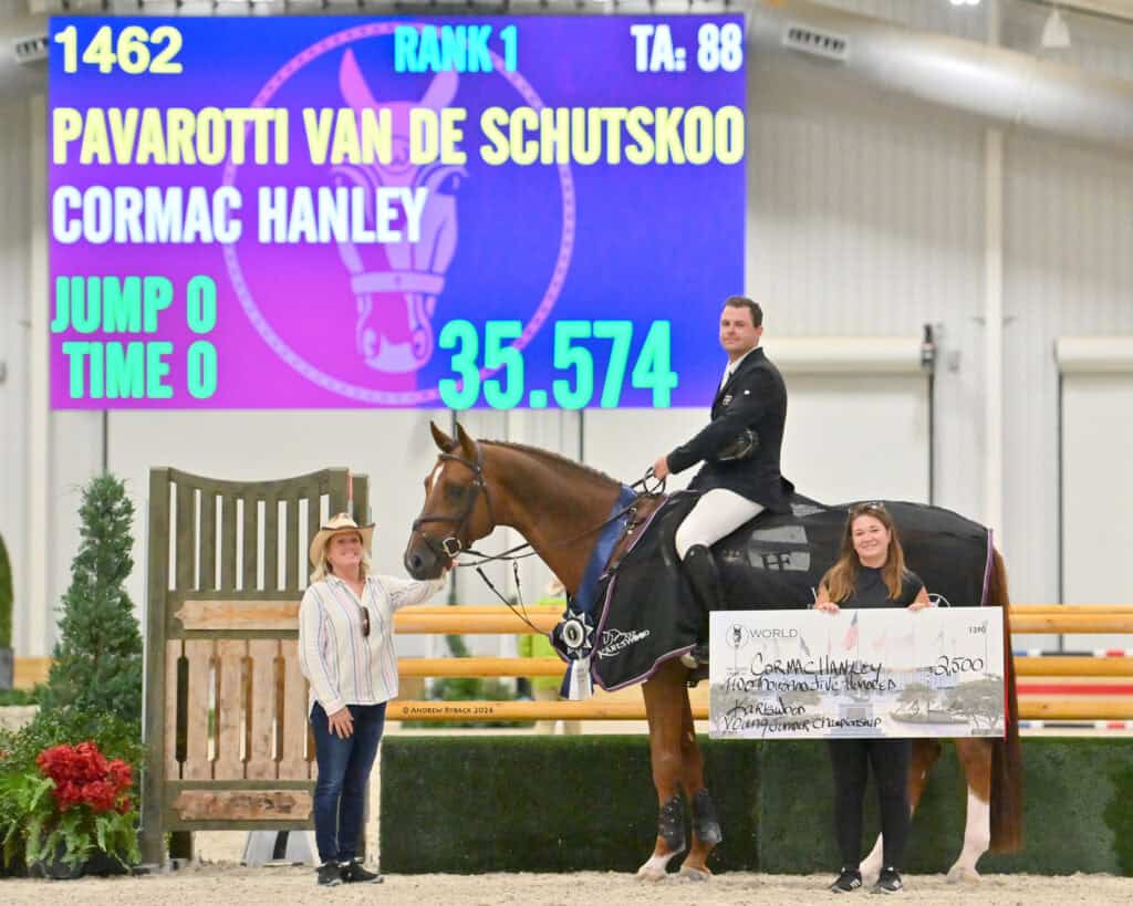 A rider on a horse poses with two women, one holding a large check. A scoreboard in the background displays rank, time, and jump details.