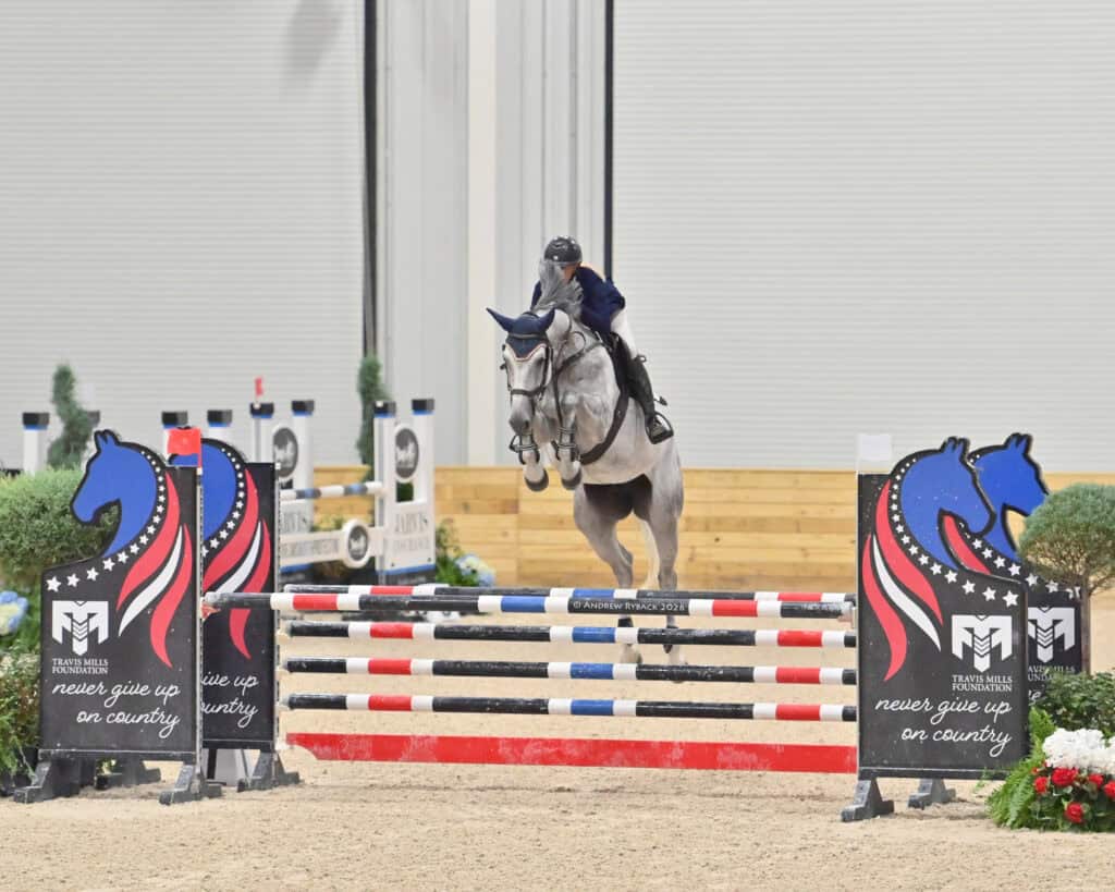 A rider in equestrian gear guides a gray horse over a show jumping obstacle with patriotic decorations in an indoor arena.