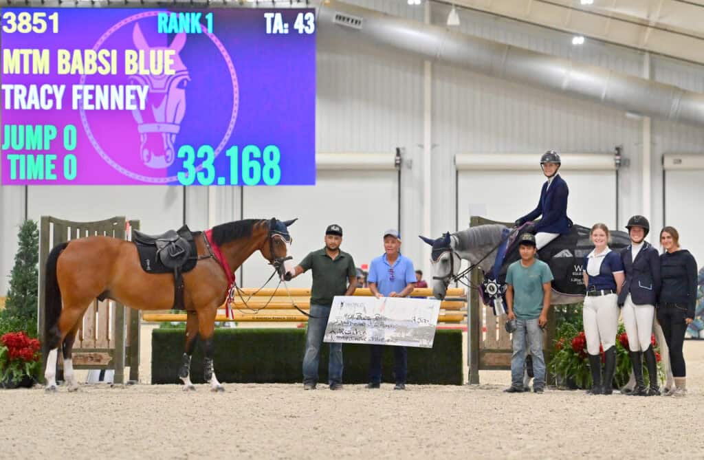 A group poses with a horse and rider in an indoor arena. A digital scoreboard behind them shows rank 1 for MTM Babsi Blue and Tracy Fenney with a time of 33.168.