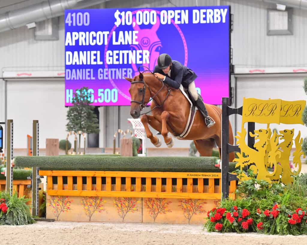 A rider in formal attire jumps a brown horse over an obstacle at an indoor event; a large screen displays derby results and names in the background.