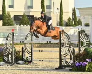 Equestrian rider in helmet and jacket jumps a brown horse over a decorative obstacle during a show jumping competition outdoors.