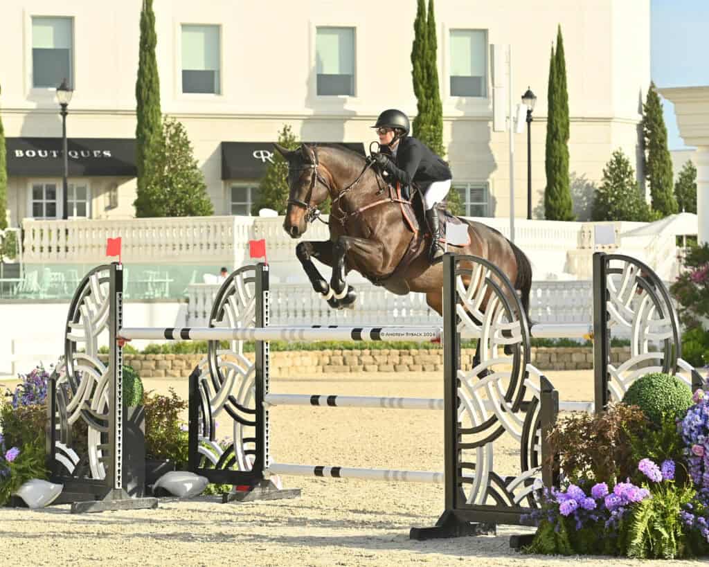 A rider in a helmet and jacket guides a horse over a jump during an equestrian event, with flowers and buildings in the background.