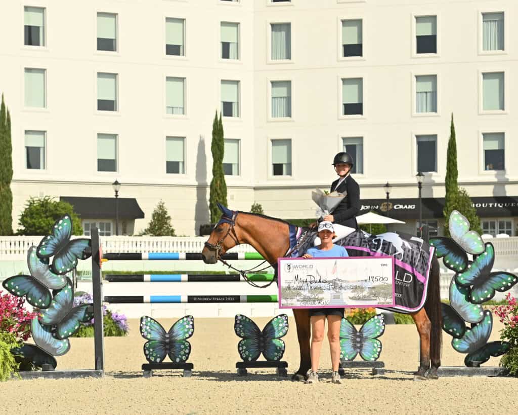A rider on a horse and a person holding a large check pose for a photo in an outdoor arena decorated with butterfly cutouts, in front of a white building.