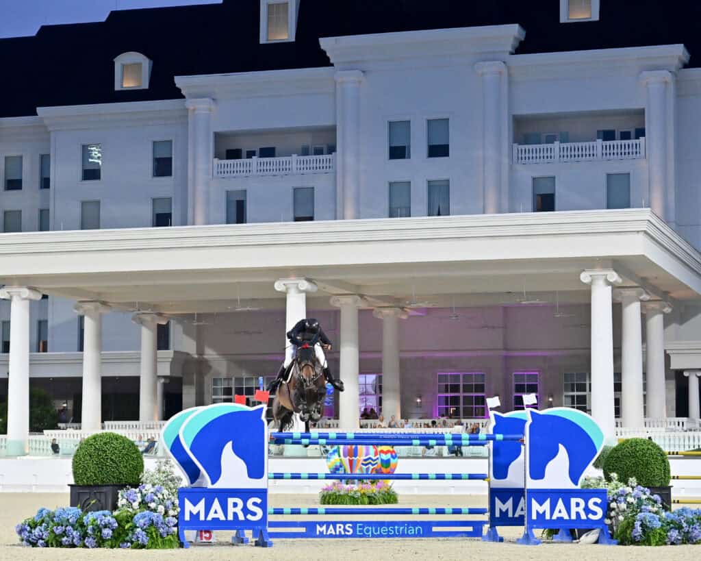 A rider on horseback jumps over a blue and white MARS-branded obstacle in front of a large, white building during an equestrian event.