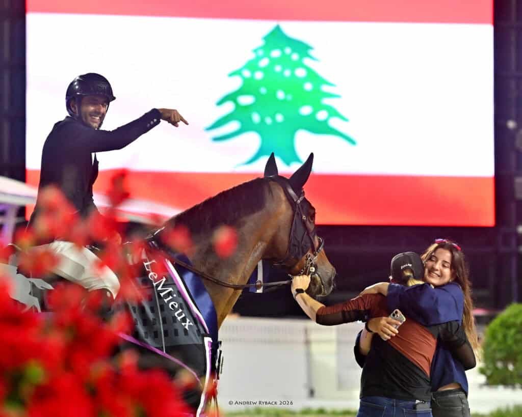 A smiling equestrian on horseback points forward as two people embrace beside the horse, with the Lebanese flag displayed in the background.
