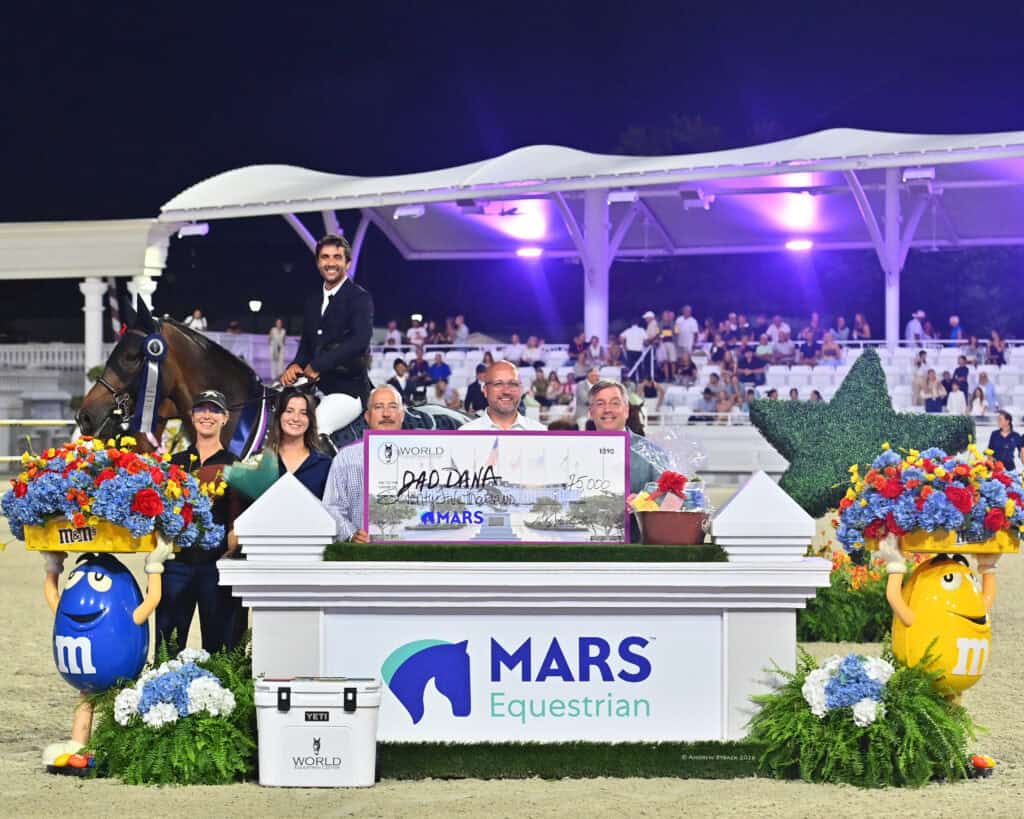 A group of people pose with a large check and floral arrangements at a MARS Equestrian event, with a rider on horseback and M&M’s mascots standing by.