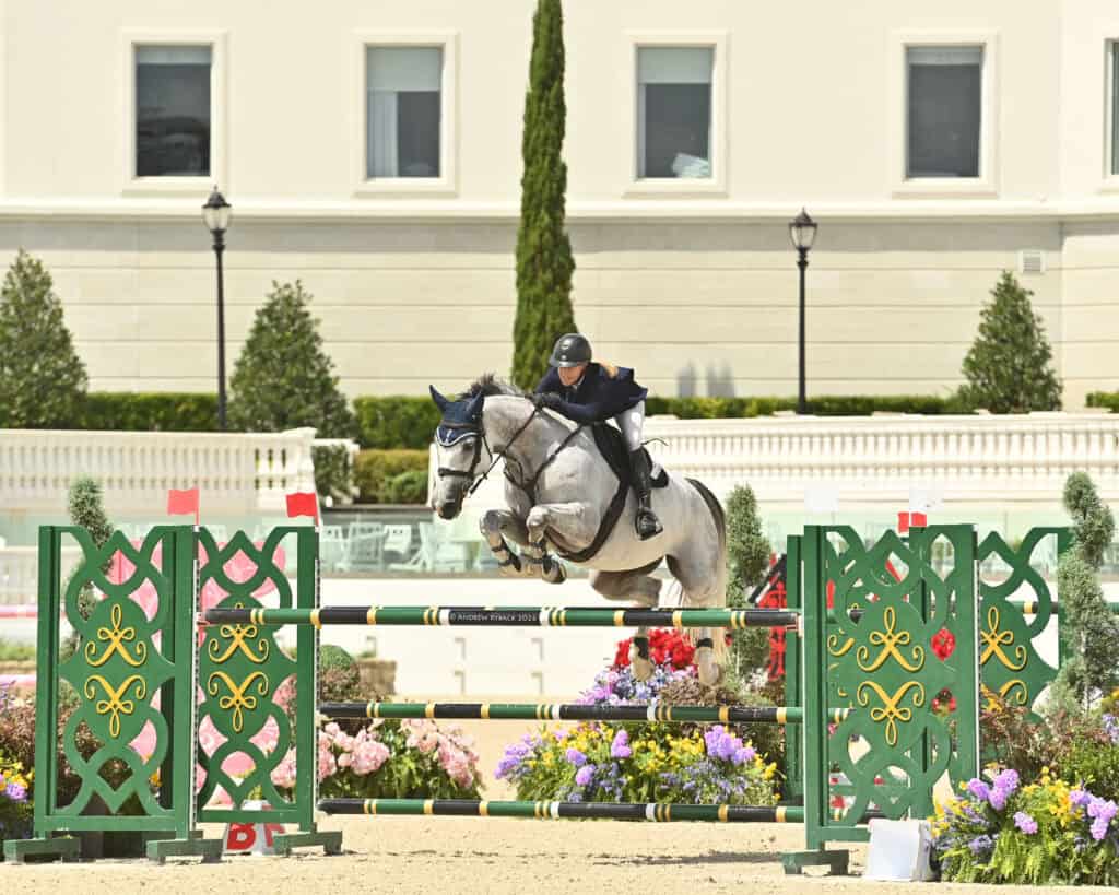 A rider wearing a helmet and dark outfit guides a gray horse over a green and yellow jump during an outdoor equestrian event.