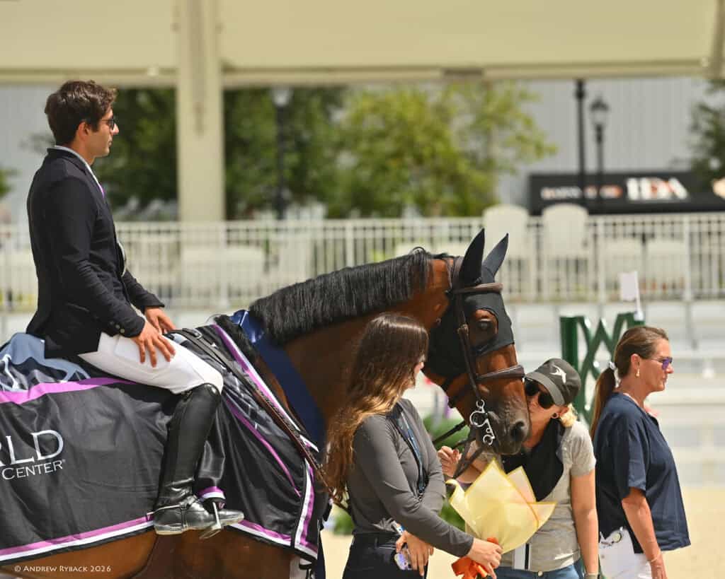 A rider in formal attire sits on a horse draped with a blanket, while three people stand nearby, one holding a yellow envelope. The background shows empty bleachers and a building.