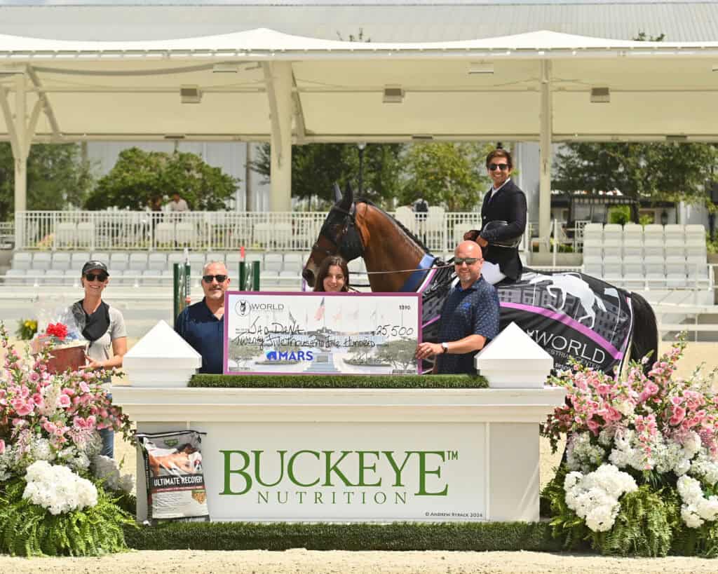 Six people and a horse pose with an oversized check for $5,500 at an outdoor equestrian event, in front of a sign that reads "Buckeye Nutrition," surrounded by flowers.