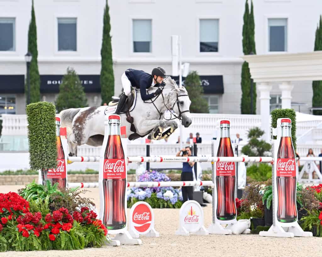 A rider on a gray horse jumps over an obstacle decorated with Coca-Cola bottles during an equestrian event.