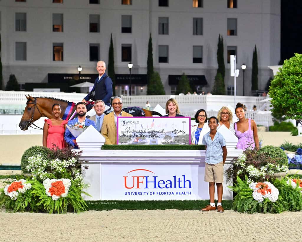 A group of people and a horse pose behind a large ceremonial check and a UF Health sign at an outdoor event, with a building in the background and flowers in the foreground.