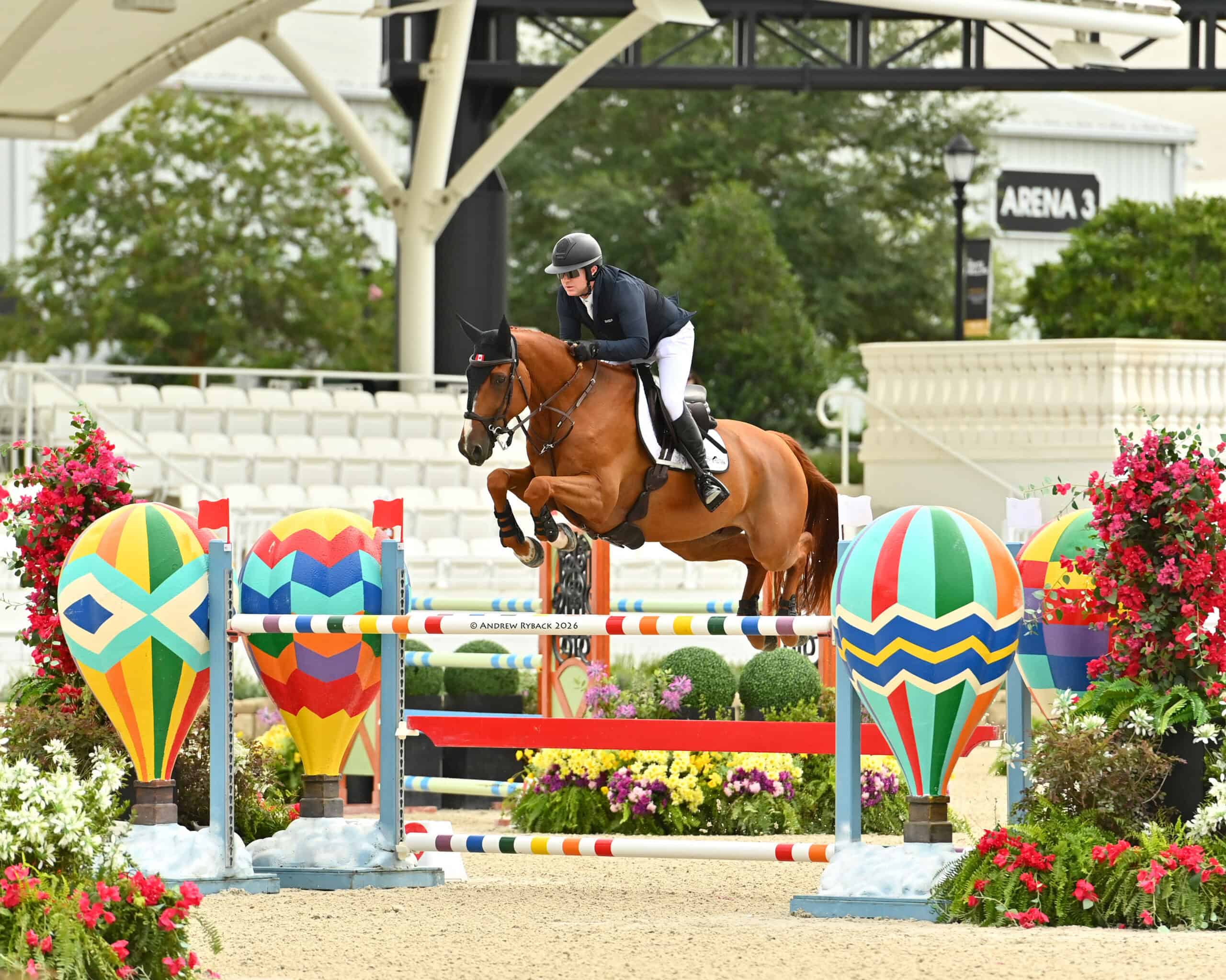 A rider on a brown horse jumps over a colorful obstacle decorated with hot air balloons in an equestrian arena.
