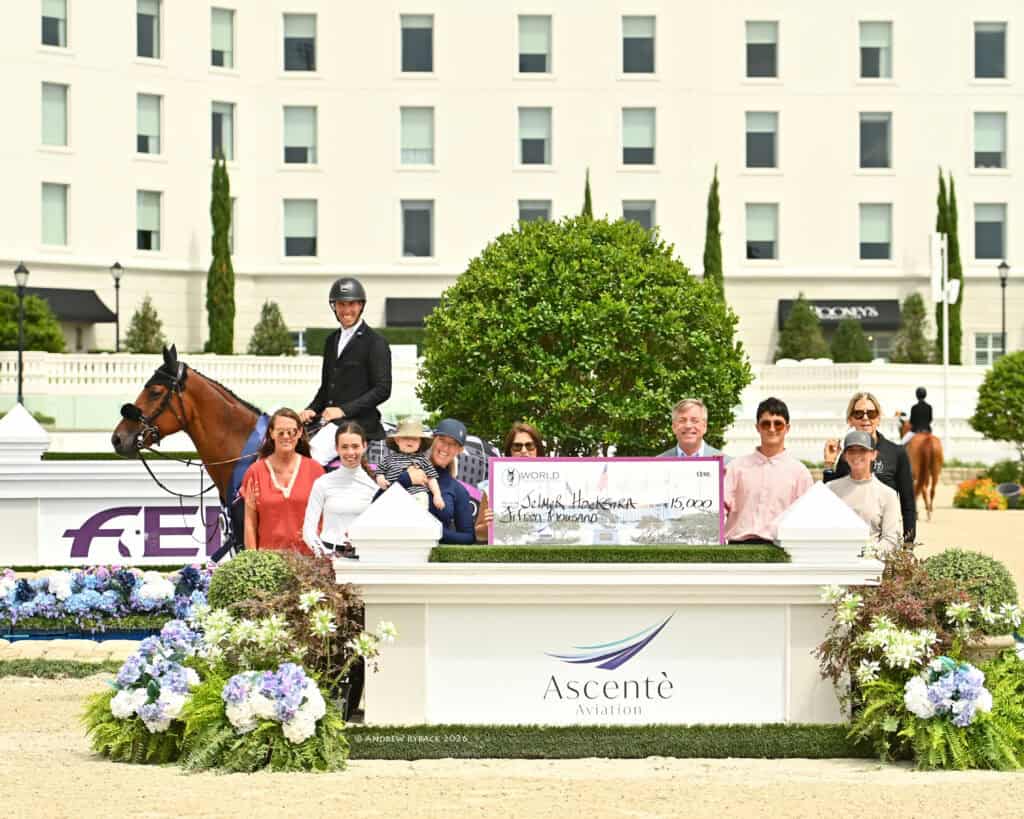A group of people and a person on horseback pose with a large check and sign reading "Ascentè Aviation" at an outdoor event, with flower arrangements in the foreground.