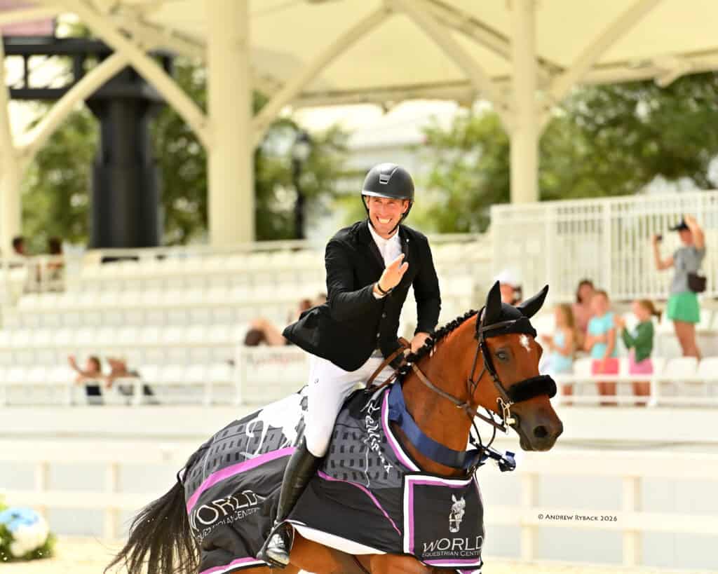 Equestrian athlete riding a horse waves while wearing a helmet and jacket at an outdoor event, with spectators in the background.