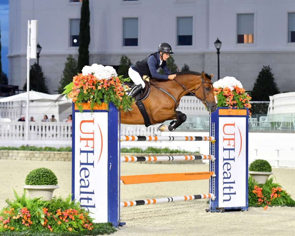 Equestrian rider wearing a helmet and vest jumps a brown horse over an obstacle with UF Health branding at an outdoor arena.