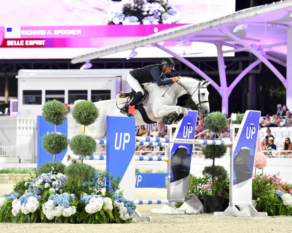 A rider on a white horse jumps over a blue and white obstacle during an equestrian event, with spectators watching in the background.