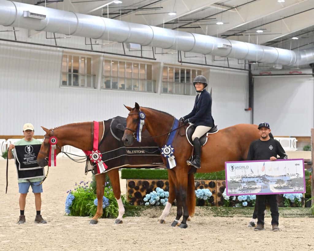 Two men and a woman with a horse inside an arena; the horse wears ribbons, the woman is riding, and one man holds a large check.