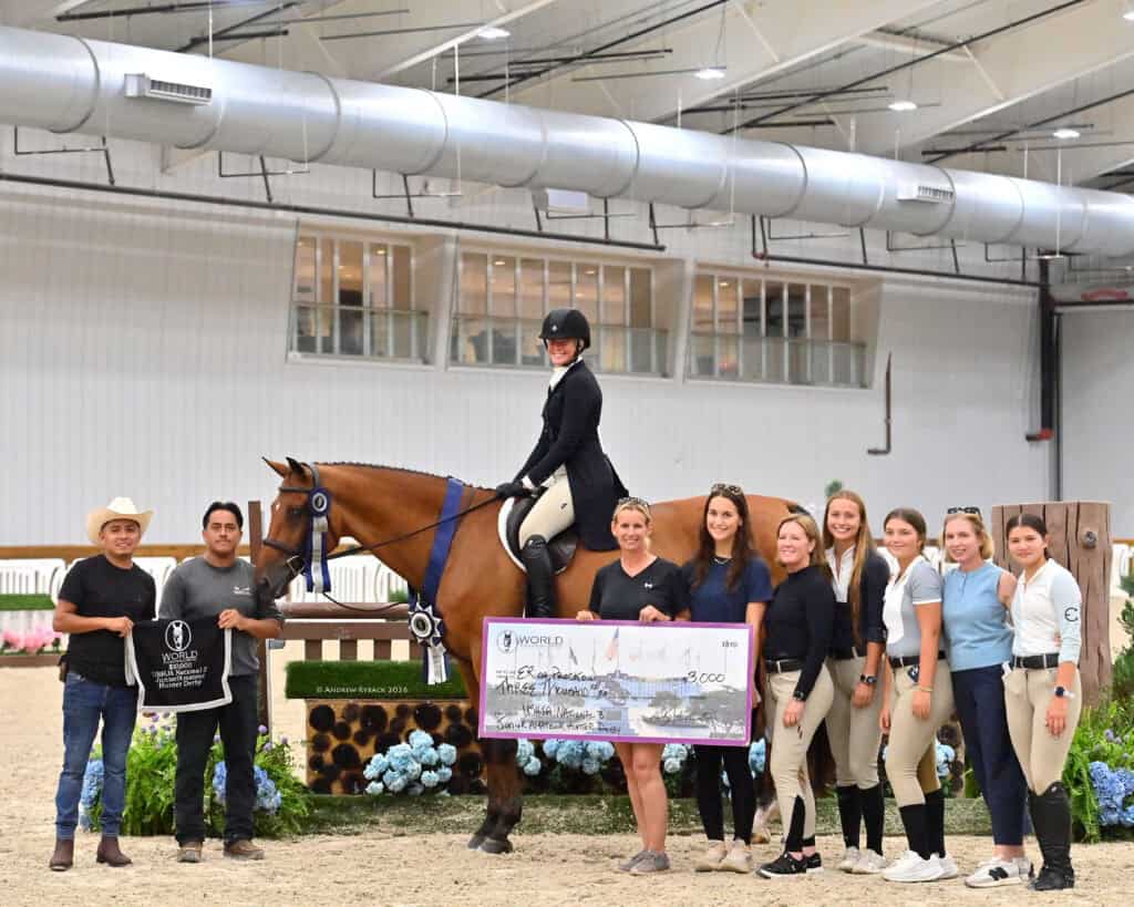 A group poses indoors with a rider on a horse, a large check, and awards after an equestrian competition.