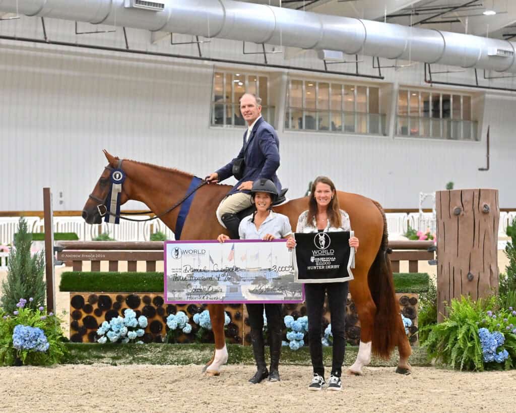 A man on a horse poses indoors while two women stand in front holding a large check and a "World Equestrian Center" sign.
