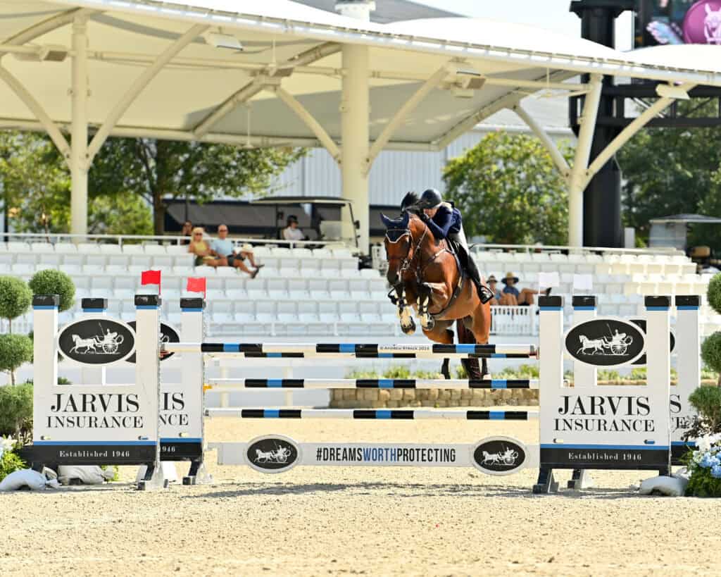 A horse and rider jump over a Jarvis Insurance-branded obstacle during an equestrian event, with white seating and spectators in the background.