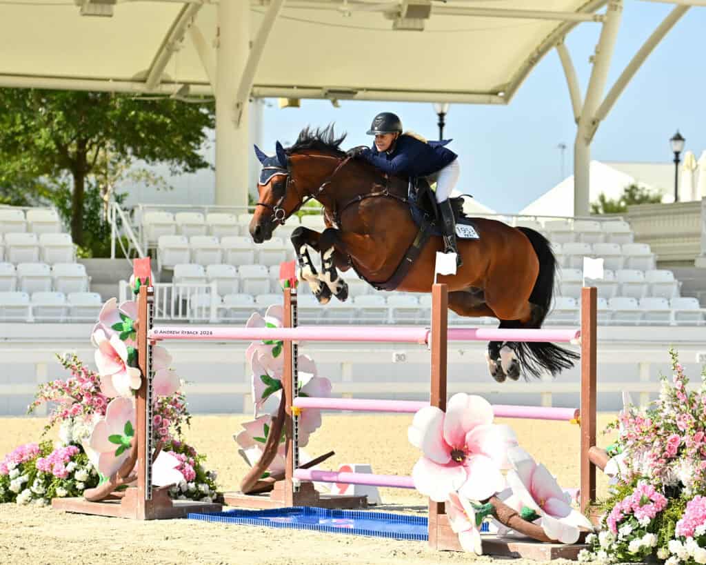 A rider in navy attire guides a brown horse over a pink and white floral jump in an empty equestrian arena.