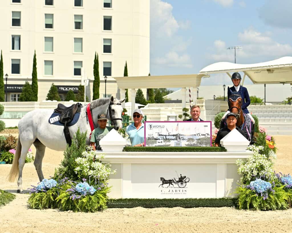 Five people and two horses pose outdoors with a large check for $15,000 in front of a sign reading "G. Jarvis" and decorative plants, with a hotel in the background.