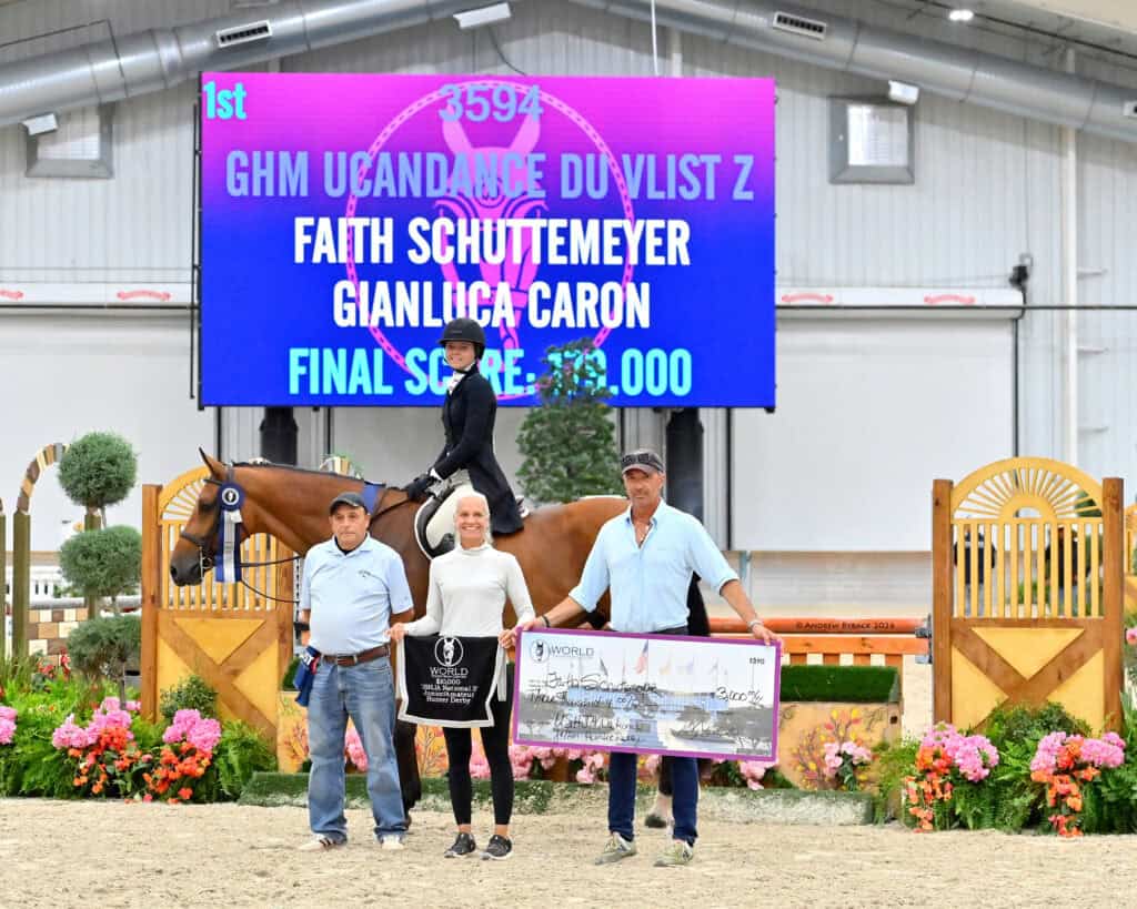 A rider on a horse poses with three adults in front of a scoreboard displaying competition results and a large prize check.
